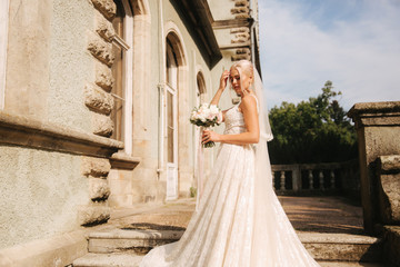 Obraz premium Gorgeous bride in wedding dress standing on the stairs by the palace. She hold a bouquet of flowers