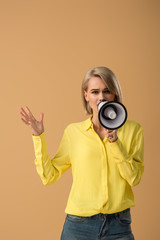Fototapeta premium Worried blonde woman in yellow shirt talking in megaphone isolated on beige