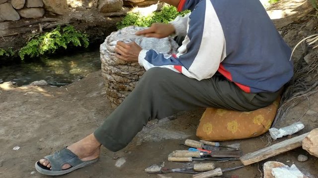 Moroccan artisan is working in the shadow of trees by the water stream, he is shaping figurine from stone using rasp tool