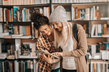Two hipster female Caucasian students standing in library and using tablet. In background shelves with books.