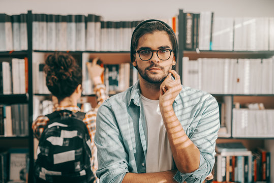 Young mixed race hipster student with eyeglasses and hat on head posing in library. In background girl taking book from shelf.