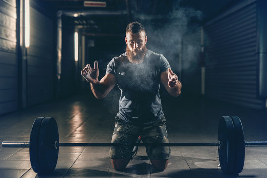 Strong muscular Caucasian bodybuilder clapping hands and crouching in hall. In front of him barbell, chalk all around. Back light, night workout concept.