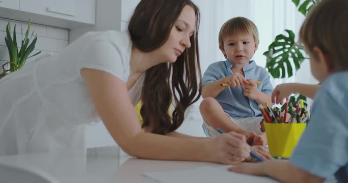 Mom helps sons learn to draw doing homework preschool preparation at home sitting in the white kitchen. Two children brothers draw a portrait of mother together