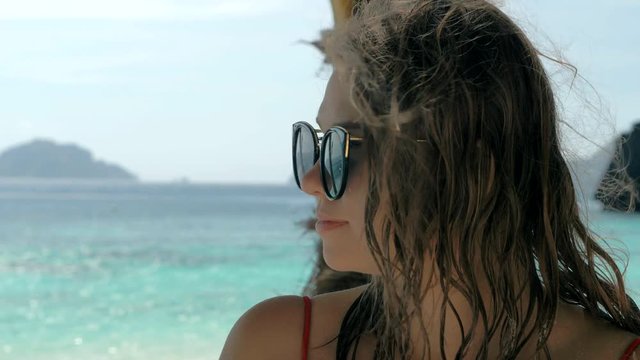 Close-up slow motion portrait of beautiful european young woman in sunglasses, hair blowing in wind on tropical beach, blue sea on the background. Travel concept.