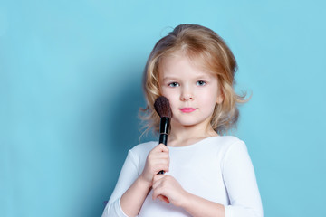 A little girl with blond hair is holding a makeup brush. Beautiful child. Photo with space for text.
