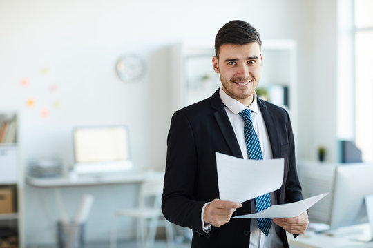 Smiling young successful banker in elegant suit looking through financial documents in office