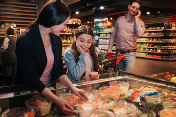Young parents and daughter in grocery store. Woman hold box of salad in hands and smile. Girl stand besides and look at it. Young man talk on phone and look at models.