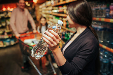 Young parents and daughter in grocery store. Woman hold water bottle and look at her family. Father and daughter stand at grocery trolley.
