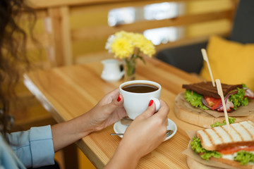 woman hold a cup of coffee at backgroud wooden table, on which lies a sandwich