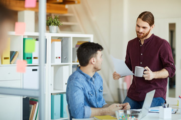 Casual young manager with mug and paper standing by desk and discussing working points with colleague