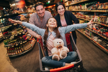 Fototapeta na wymiar Young parents and daughter in grocery store. Playful girl has toy bear on knees. She pretend flying. Parents standing behind and push trolley with girl.