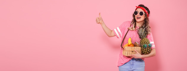 Beautiful young girl in pink t-shirt and glasses, holds a full straw bag of fruit on pink background. Shows finger class
