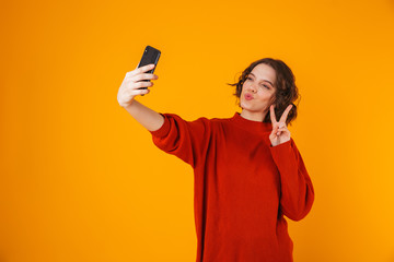 Portrait of beautiful woman holding and taking selfie photo on cell phone while standing isolated over yellow background