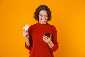 Portrait of brunette woman using cell phone and credit card while standing isolated over yellow background