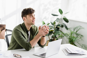 handsome businessman in green shirt sitting, holding cup near table with laptop in office
