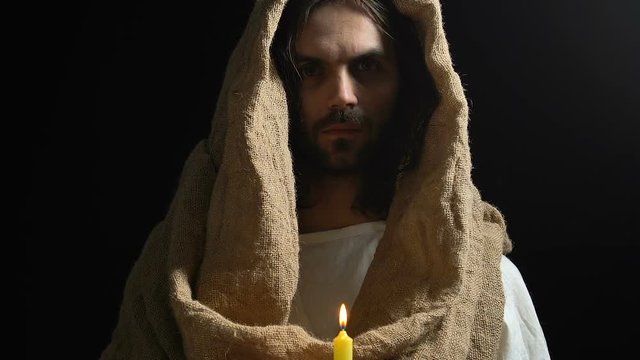 Jesus Christ in robe holding burning candle against dark background, light