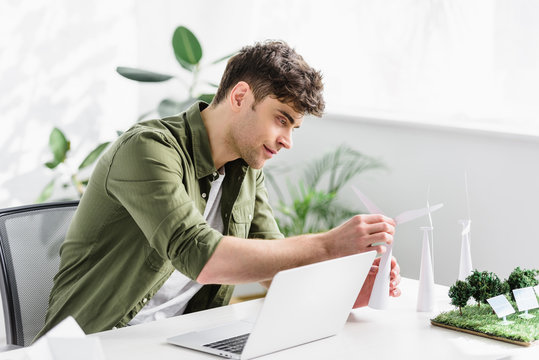 handsome architect sitting at table with laptop and putting windmill model in office