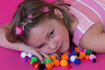 Horizontal closeup of beautiful young girl in pink, orange and white dress lying down on colourful pom poms 