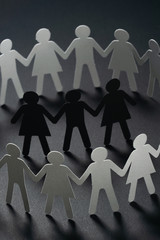 Three human paper figures surrounded by circle of paper people holding hands on dark surface....