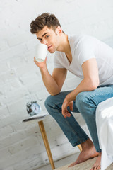 Fototapeta na wymiar man in white t-shirt and jeans sitting on bed and drinking coffee in bedroom