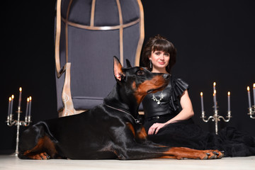 A girl in a black dress is sitting on the floor next to a gray chair and a Doberman dog is next to...