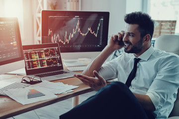 Great results! Young businessman in formalwear talking on the smart phone and smiling while sitting in the office