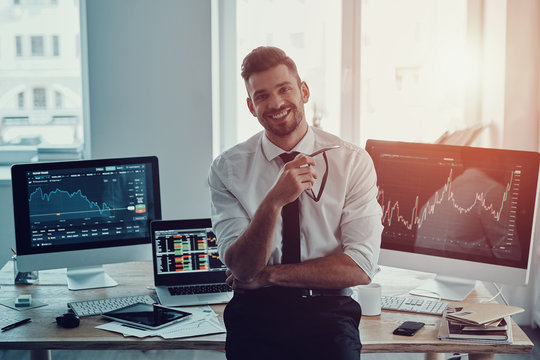 Happy businessman. Handsome young man in formalwear keeping hand on chin and looking at camera with smile while standing in the office - Powered by Adobe