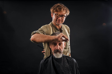 Hairdresser cuts senior citizen with a beard on a dark background in the studio