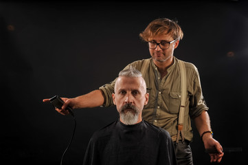 Hairdresser cuts senior citizen with a beard on a dark background in the studio