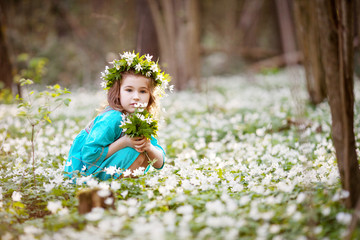 Beautiful little girl in a blue dress walking in the spring wood. Portrait of the pretty girl with a wreath from flowers on the head. 