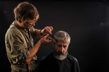 Hairdresser cuts senior citizen with a beard on a dark background