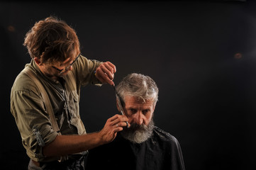 Hairdresser cuts senior citizen with a beard on a dark background