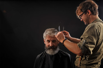 Hairdresser cuts senior citizen with a beard on a dark background