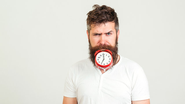 Handsome man holds the clock with his mouth. Crazy bearded hipster with clock. Brutal bearded guy with alarm clock on white background. People and time concept.
