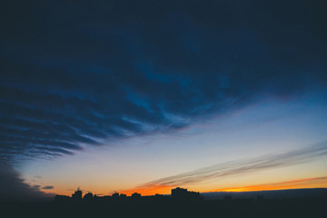 Cityscape with wonderful varicolored vivid dawn. Amazing dramatic blue cloud sky above dark silhouettes of city building roofs. Atmospheric background of orange sunrise in overcast weather. Copy space