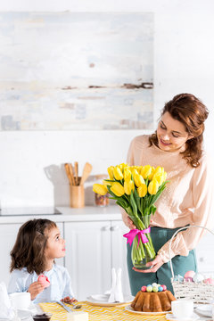 Child eating macaroon and looking at mother with tulips bouquet in kitchen