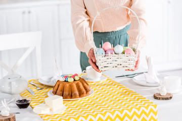 Cropped view of woman holding wicker basket with painted easter eggs near table