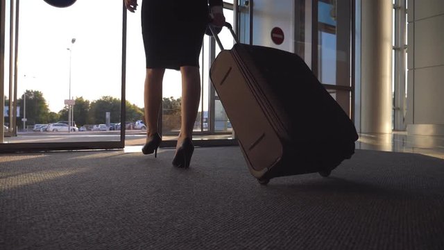 Business lady leaving airport through automatic glass door with her luggage. Young woman in heels walking from terminal and roll suitcase on wheels. Concept of work trip or travel. Slow motion