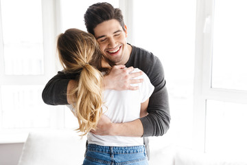 Portrait of joyful couple smiling and hugging together while standing near big window at home
