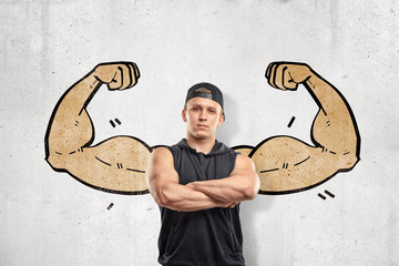 Close up front view of young athletic man with arms crossed, standing against wall with drawing of big muscular arms right from behind his back.