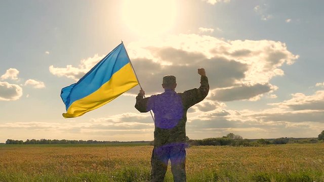 Soldier stand with  Ukrainian flag and hand up against evening sky with sun. Outdoor slow motion scene