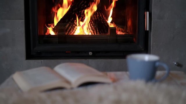 Hygge concept with open book and cup of tea near burning fireplace