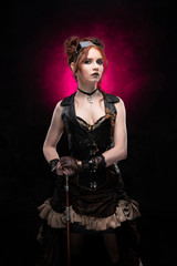 A beautiful red-haired cosplayer girl wearing a Victorian-style steampunk costume with large breasts in a deep neckline and glasses holding a cane on a black and purple background