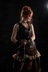 A beautiful red-haired cosplayer girl wearing a Victorian-style steampunk costume with a big breast in a deep neckline stands thoughtfully, leaning on a cane, on a dark background.