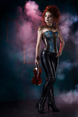 Beautiful redhead cosplayer girl wearing a steampunk corset and leather pants with a big breast holds violin in her hand in theatrical smoke.