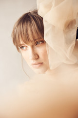 Soft beauty portrait of young dreamy woman with blue eyes and bang, weared in nude tulle dress. Studio shot