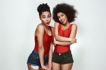 Pure beauty. Portrait of two cute and shy afro american women in jeans shorts are making a face while standing against grey background