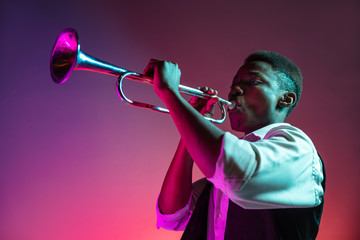 African American handsome jazz musician playing trumpet in the studio on a neon background. Music concept. Young joyful attractive guy improvising. Close-up retro portrait. - obrazy, fototapety, plakaty