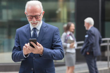 Successful happy senior gray haired businessman using smart phone, browsing internet or messaging. Two people talking in the background, blurred