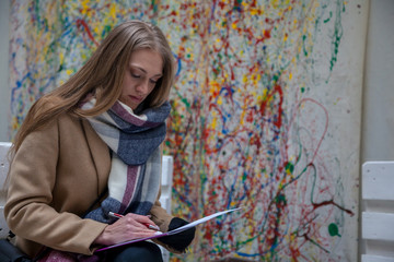 Young blonde woman siting on bench and writing. Woman in coat and scarf writing outdoor.